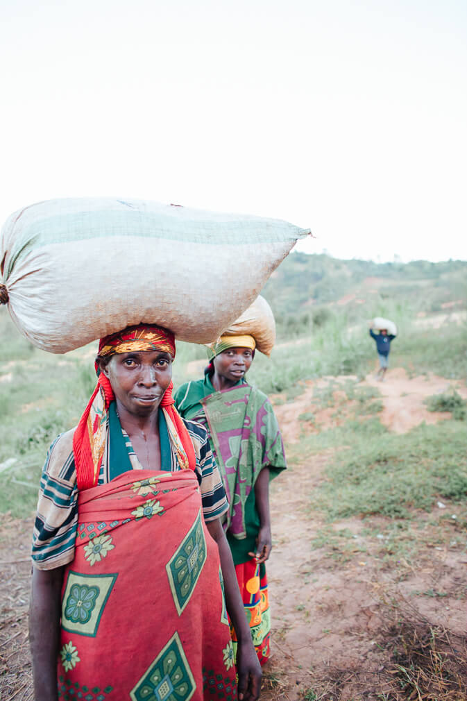 Two Burundian women carrying sacks of coffee on their heads while standing on a dirt path